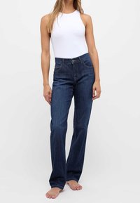 Jean oscuro con un diseño de corte recto, bolsillos delanteros y cinturilla. Combinado con un bodysuit sin mangas de color blanco. No se ven zapatos.