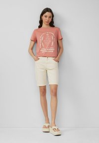 Różowa koszulka z białym wzorem graficznym, w połączeniu z jasnobezowymi szortami. Model nosi sandały na platformie z paskami. Proste tło.
