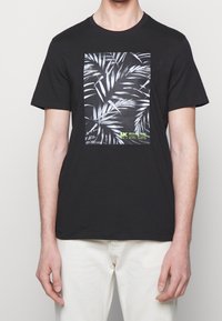 Hombre vestido con una camiseta negra con un gráfico de hoja de palma blanca y un pequeño texto amarillo, acompañado de pantalones blancos, de pie contra un fondo gris claro.
