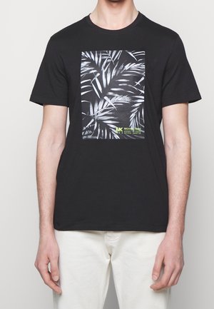 Mann trägt ein schwarzes T-Shirt mit weißem Palmblattgrafik und kleinem gelbem Text, kombiniert mit weißen Hosen, steht vor einem hellgrauen Hintergrund.