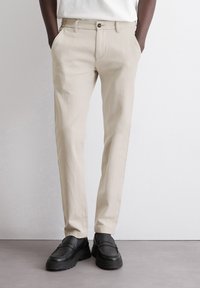 Beige trousers with a slim fit, straight leg design, featuring a front button closure and side pockets. Worn with black shoes.