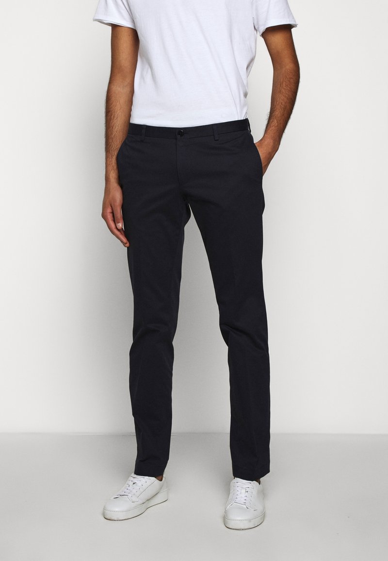 Homme portant un pantalon slim noir, un t-shirt blanc et des baskets blanches, debout avec une main dans la poche devant un fond uni.