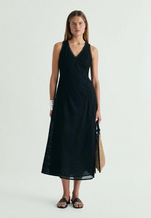Joven mujer de pie con un vestido midi negro sin mangas con detalles de encaje, sandalias negras, pulsera plateada, y sosteniendo un bolso tejido.