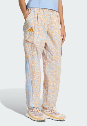 Person iført lyseblå og orange leopardmønstrede træningsbukser med Adidas-logo og matchende sneakers, stående med hænderne i lommerne.