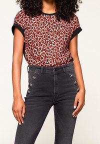 Rot-schwarz gemusterte Kurzarmbluse mit umgeschlagenen Ärmeln, kombiniert mit hochgeschnittenen dunklen Jeans mit dekorativen seitlichen Knöpfen.