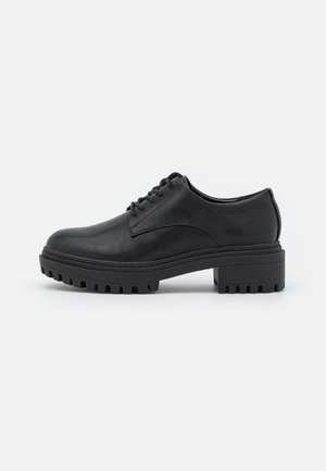 Chaussure noire en cuir à lacets avec un bout rond, semelle en caoutchouc striée et solide talon carré, présentant une texture lisse et un design minimaliste.
