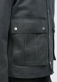 Close-up af mørkegrå jakkelomme med klap sikret med to sølvfarvede trykknapper på siden af en person.