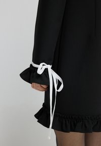 Černé šaty s nařasenými manžetami, které mají bílé stužky. Volánkový lem dodává texturu. Tkanina vypadá hladce a lehce.