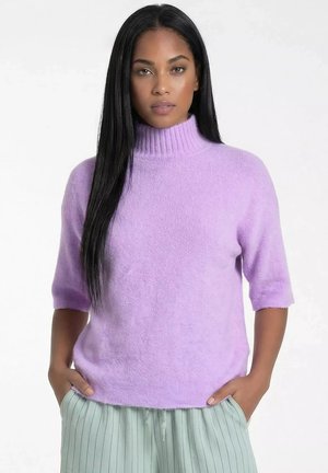 Kobieta z długimi czarnymi włosami, ubrana w lawendowy sweter z krótkim rękawem i golfem oraz jasnozielone spodnie w paski, z rękami w kieszeniach.