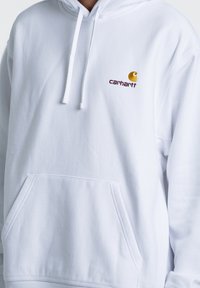 Sudadera blanca con bolsillo frontal. Presenta una capucha con cordón y un logo de Carhartt bordado en amarillo y marrón en el lado izquierdo del pecho.