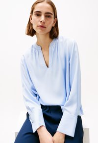 Blusa azzurra chiara con scollo a V e colletto arricciato. Maniche lunghe con polsini larghi, realizzata in tessuto morbido, abbinata a pantaloni blu navy.