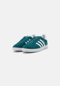 Teal suede sneakers with white leather accents and three stripes. Round toe and textured rubber sole. Features brand logo in gold.