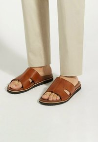 Bruine leren slide sandalen met een grote opening aan de voorkant en een platte zool, met een gestructureerde afwerking en minimale ontwerpaccenten.