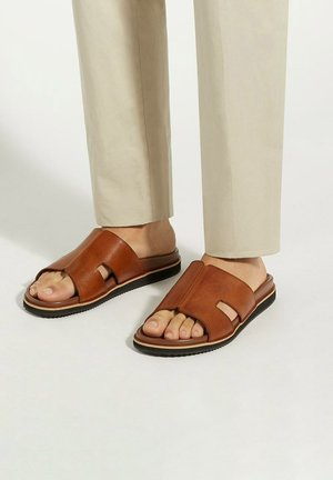 Sandales à glissière en cuir marron avec une grande ouverture à l'avant et une semelle plate, présentant une finition texturée et des accents de design minimal.