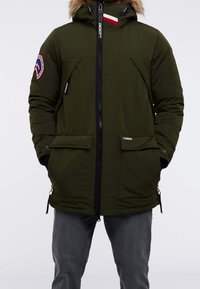 Green padded winter jacket with a fur-trimmed hood, front zipper, two large pockets, and logo patch on the sleeve.