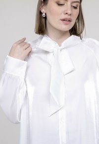 Blouse blanche avec un éclat satiné, présentant un col à nœud, un plastron de boutons et des manches longues légèrement bouffantes.
