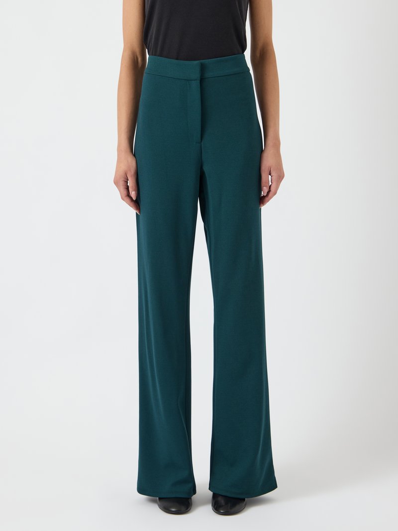 Tegen de groene wijde benen broek gemaakt van een gladde stof met een hoge taille en een rits aan de voorkant. Heeft een schoon ontwerp en minimale hardware.