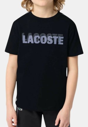 Bambino che indossa una maglietta nera a maniche corte con il logo "LACOSTE" pixelato color viola chiaro sul petto.