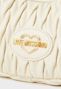 Bež usnjena torbica s quilted dizajnom, ki ima okroglo oznako z napisom "LOVE MOSCHINO" v zlatih črkah.