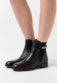 Bottines en cuir noir avec des talons bas, portées sur des jambes nues, dotées de panneaux élastiques sur les côtés et d'un détail en or "HILFIGER" sur la sangle.
