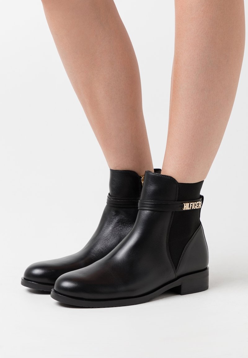 Bottines en cuir noir avec des talons bas, portées sur des jambes nues, dotées de panneaux élastiques sur les côtés et d'un détail en or "HILFIGER" sur la sangle.