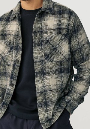 Homme portant une chemise en flanelle beige et noire à motifs, boutonnée, par-dessus un t-shirt noir, avec une main dans la poche, sur fond uni.
