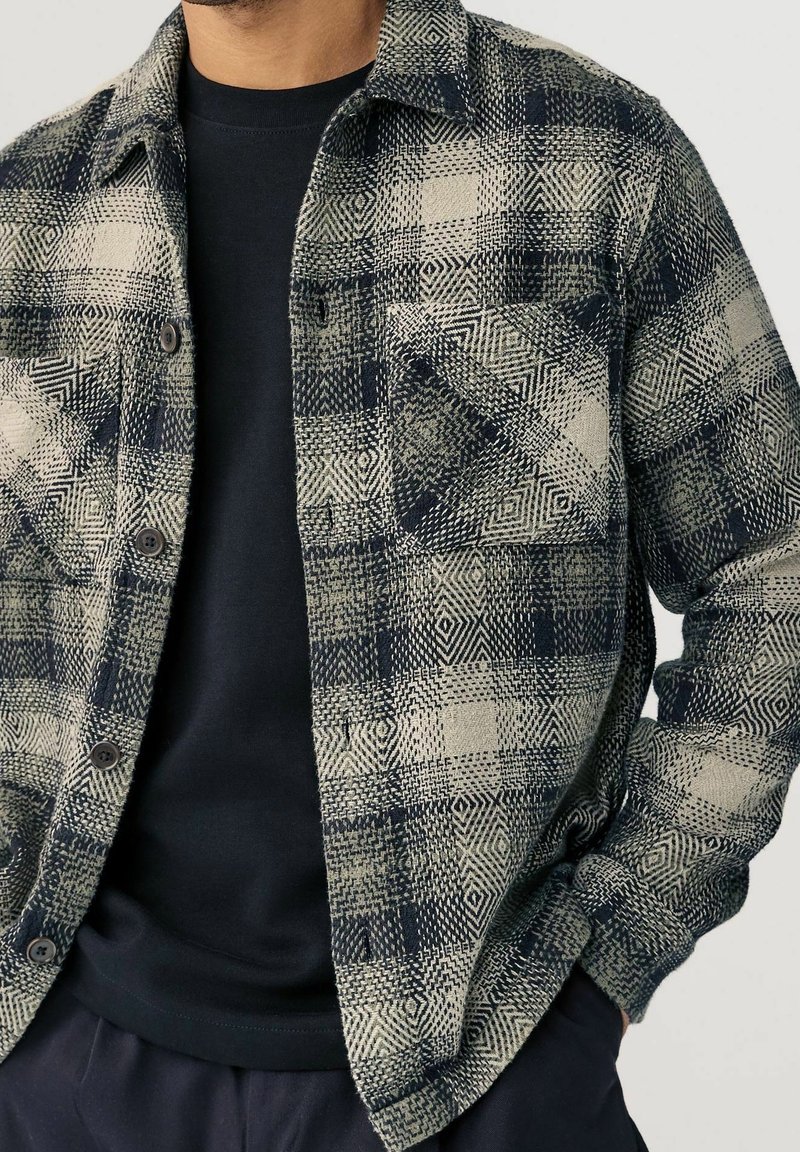 Homme portant une chemise en flanelle beige et noire à motifs, boutonnée, par-dessus un t-shirt noir, avec une main dans la poche, sur fond uni.