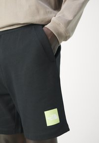 Pantalones cortos de algodón negros con bolsillos laterales y un parche cuadrado verde claro que presenta el texto y emblema de "The North Face".