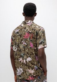 Ein Mann, der von hinten zu sehen ist und ein kurzärmliges braunes Hemd mit einem floralem Muster in Pink, Lila und Weiß trägt, vor einem schlichten, hellen Hintergrund.