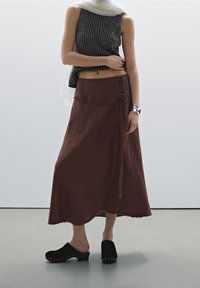 Nainen, jolla on yllään hihaton harmaa raidoitettu toppi, pitkä ruskea kietaisuhame, jossa on solkia, mustat puuvarsikengät ja hopeaketju, seisoo sisätiloissa.