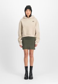 Beige krāsas īsais džemperis ar priekšējo kabatu, saskaņots ar zaļu mini svārku, papildināts ar melniem ceļgala zābakiem un melnu cepuri.