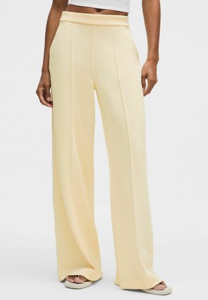 Nainen pukeutuneena vaaleankeltaisiin korkeavyötäröisiin leveälahkeisiin housuihin, joissa on etusaumat, ja valkoisiin avokkaisiin, seisoo yksivärisen taustan edessä.