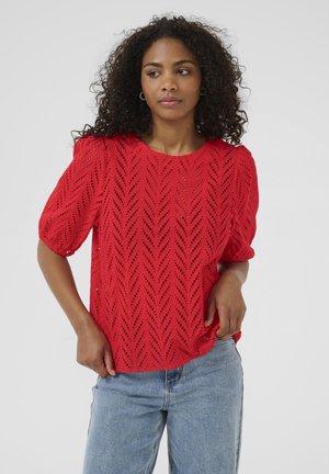 Blouse rouge à manches courtes avec un motif de feuilles texturé et des découpes ouvertes. Col rond et coupe décontractée, associée à un jean bleu clair.