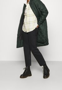 Lungo cappotto imbottito verde sopra una camicia a quadri chiari con accenti gialli, abbinato a pantaloni neri slim-fit e stivali neri stringati.