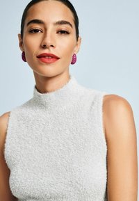 Mouwloze top met hoge hals in zacht, gestructureerd lichtgrijs stof; gecombineerd met gedurfde, gestructureerde fuchsia oorbellen. Gladde huid en rode lippen.