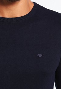 Pull en maille bleu marine avec un col ras du cou. Logo discret brodé sur la poitrine. Le tissu semble doux avec une texture lisse.