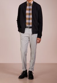 Giubbotto bomber nero, camicia a quadri nei toni marroni/grigi, pantaloni grigi chiari a forma affusolata e stivali neri con dettaglio delle stringhe.
