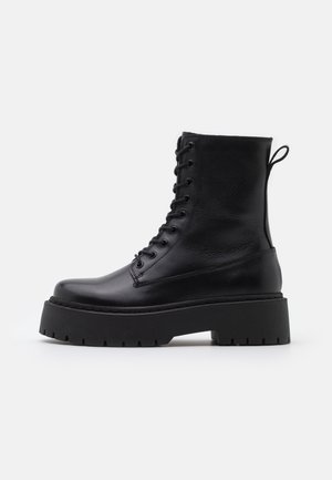 Botte de combat en cuir noir à lacets avec semelle épaisse à crampons et languette arrière sur fond blanc uni.