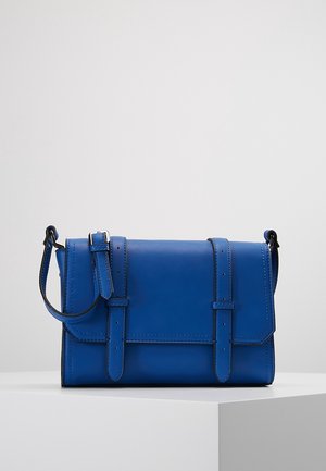 Blå læder crossbody taske med frontklaff, der er sikret med to spændestropper, placeret på en hvid sokkel mod en ensartet baggrund.