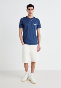 T-shirt en coton bleu marine avec le logo "Lee" sur la poitrine, associé à un short de couleur crème et des baskets blanches avec des semelles en gomme.