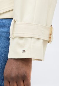 Κοντινό ενός σκούρου δέρματος χεριού που φοράει μανίκι μπεζ με μικρό μεταλλικό λογότυπο και ορατό μπλε τζιν παντελόνι.