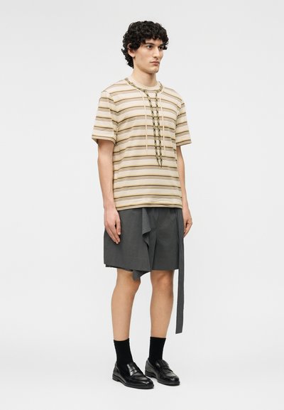 Jeune homme portant un t-shirt beige rayé avec un détail en corde, un short asymétrique gris, des chaussettes noires et des chaussures en cuir noir, debout devant un fond blanc.