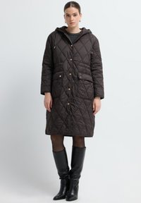 Bruine gewatteerde jas met een capuchon, met diagonale stiksels, twee voorzakken en drukknoopsluitingen. Gedragen met zwarte kniehoge laarzen.