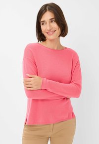 Rosa Langarmstrickpullover mit Rundhalsausschnitt und seitlichen Schlitzen. Weicher Griff mit gerippten Bündchen, lässig kombiniert mit beigen Hosen.