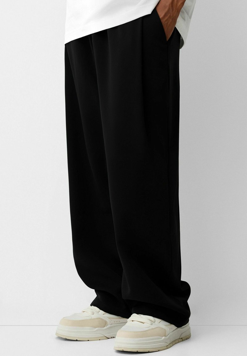 Czarne, luźne spodnie wykonane z gładkiego materiału, z bocznymi kieszeniami, w połączeniu z jasnymi sneakersami.