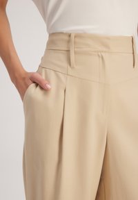 Beige op maat gemaakte broeken met een hoge taille, geplooide voorkant en zakken aan de zijkant. Gemaakt van een gladde, gemerceriseerde stof, ontworpen voor een gestroomlijnde pasvorm.
