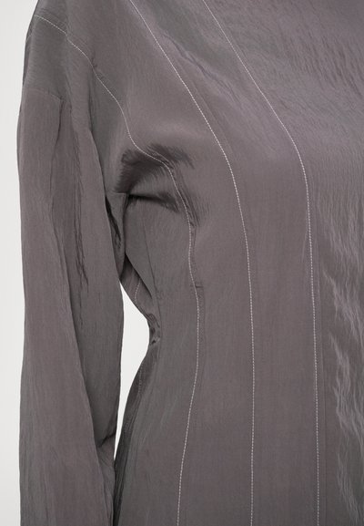 Camicia in tessuto grigio testurizzato con maniche lunghe, caratterizzata da cuciture bianche a contrasto e una vestibilità rilassata, che mette in evidenza linee di design sottili.