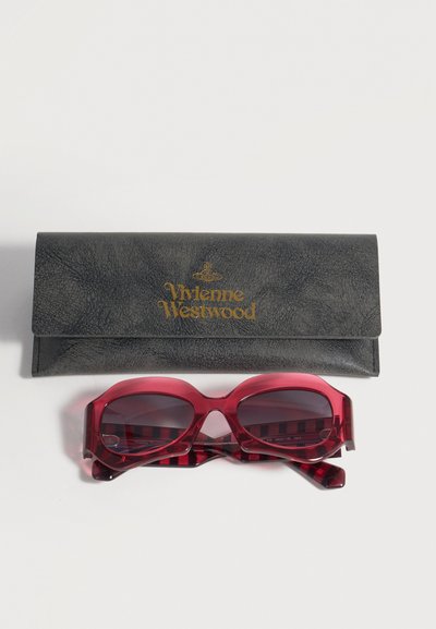 Διπλωμένη μαύρη θήκη γυαλιών Vivienne Westwood με χρυσό λογότυπο πάνω από ένα ζευγάρι διαφανή κόκκινα οβάλ γυαλιά ηλίου με ριγωτούς βραχίονες.