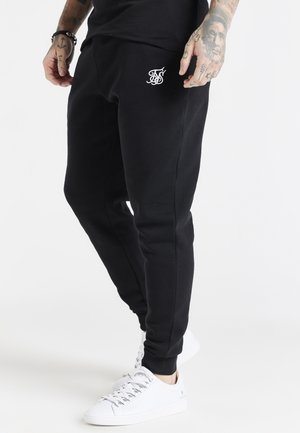 Homme portant un jogger slim noir avec un logo blanc sur la cuisse et des baskets blanches, mains et poignets tatoués visibles.