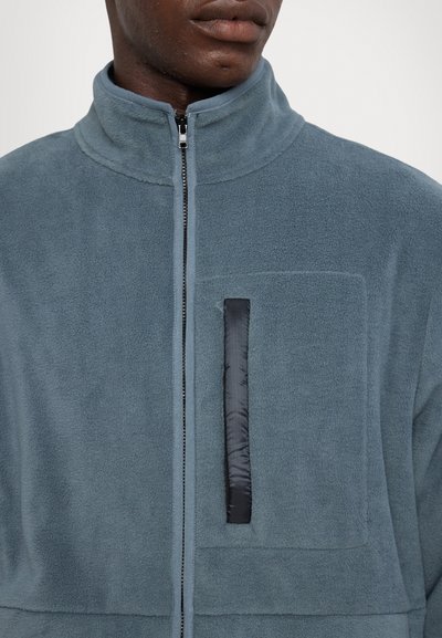 Veste en polaire bleu-gris atténué avec un col haut, fermeture éclair sur toute la longueur à l'avant, et une poche en tissu noire sur la poitrine. Texture douce et design décontracté.